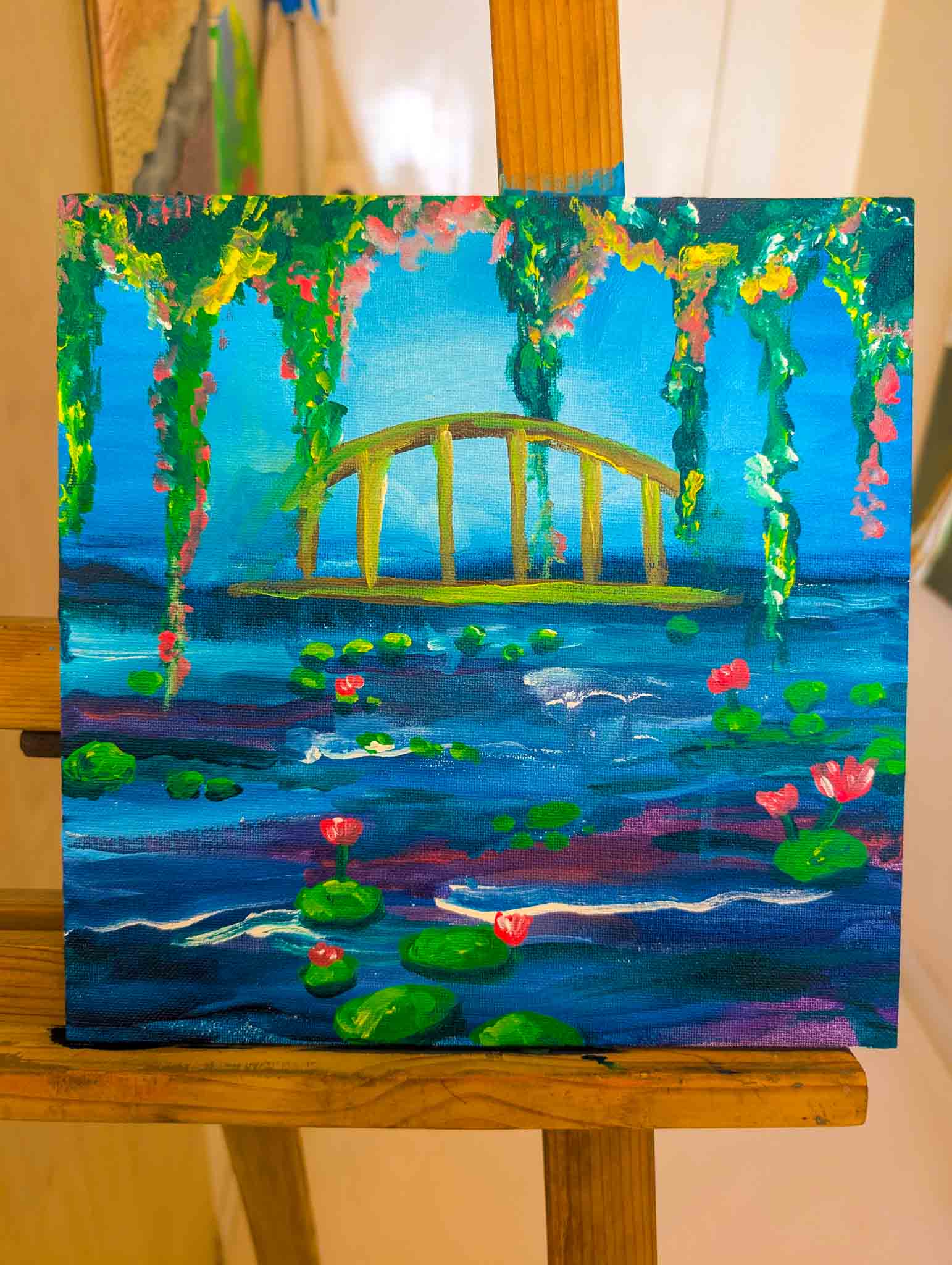 Woman holding up her colorful Sip & Paint artwork of a blue bridge and lotus-filled pond, smiling at the camera.